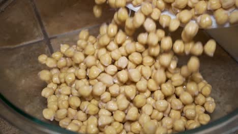 Pouring-washed-and-soaked-chickpeas-or-garbanzos-in-glass-bowl,-Overhead-Slowmo