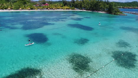 Vista-Aérea-En-Cámara-Lenta-De-Personas-Haciendo-Paddleboarding-Cerca-De-La-Playa-De-Un-Resort-En-El-Mar-Caribe-Hondureño