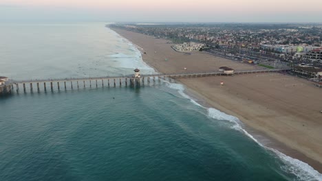 4k-ariel-drone-shot-heading-straight-down-the-pier-in-California,-Surf-City-USA-as-a-military-troop-trains,-surfers-catch-waves,-a-lifeguard-truck-drives-by-and-families-enjoy-summer-vacation