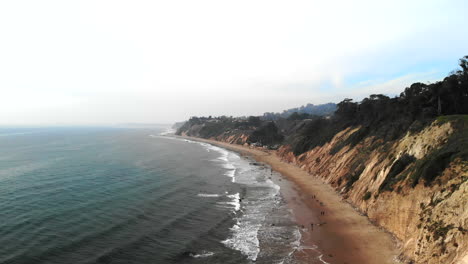 Aerial-drone-rising-over-the-blue-pacific-ocean-waves-off-the-beach-cliffs-on-the-Santa-Barbara,-California-overcast-coastline
