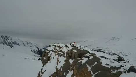 Drone-Volando-Sobre-La-Cresta-De-La-Montaña-Rocosa-Con-Un-Alpinista-De-Esquí-Solitario-Parado-En-La-Cima