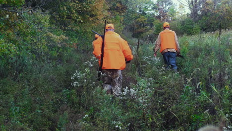 Three-men-in-blaze-orange-carrying-shotguns-walk-through-the-bush-with-their-hunting-dogs