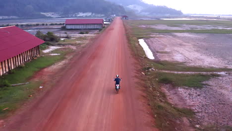 Die-Drohne-Folgt-Dem-Motorrad-Auf-Der-Kambodschanischen-Unbefestigten-Straße,-Bis-Am-Horizont-Berge-Auftauchen