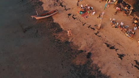 Aerial-flight-back-from-against-people-sitting-at-beach-bar-on-tropical-island-during-sunset