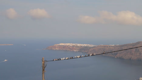 The-village-of-Oia-built-on-the-edge-of-dark-cliffs,-as-seen-from-far-away-with-pigeons-sitting-on-a-telephone-wire-in-the-foreground