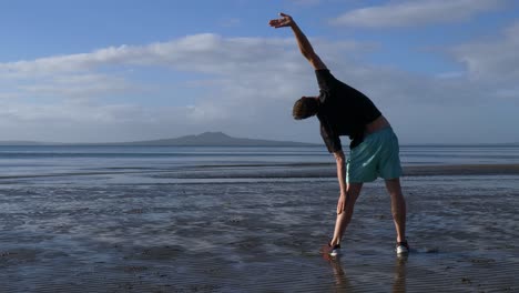 Young-fit-man-stretching-on-the-beach-in-Auckland,-New-Zealand-in-the-morning-with-Rangitoto-island-in-the-distance