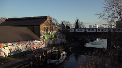 Großbritannien,-Februar-2019-–-Ein-Zug-überquert-Eine-Brücke-über-Den-Regents-Canal-Und-Eine-Mit-Graffiti-Verzierte-Wand-Auf-Einem-Treidelpfad-Mit-Dem-Finanzviertel-Canary-Wharf-Im-Hintergrund