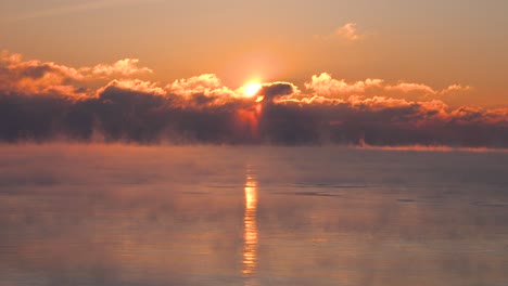 bright-yellow-red-and-orange-sunrise-through-dark-clouds-over-a-very-cold-lake-as-steam-rises-in-the-middle-of-winter