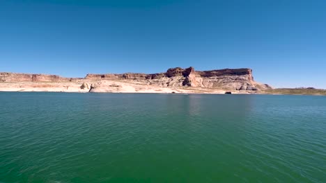 As-viewed-from-out-in-the-turquoise-waters-of-Lake-Powell,-Page,-Arizona-a-speedboat-in-the-distance-races-past-the-sandstone-buttes
