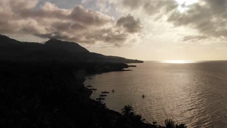 4k-drone-aerial-over-an-exotic-tropical-island-paradise-in-the-Pacific-Ocean-at-dusk-and-sun-rays-shining-brightly-through-a-cloudy-sky-and-volcano-peak-and-dark-coastline-with-boats-on-the-sea