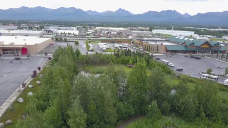 Aerial-footage-of-a-pond-at-a-Cabela's-parking-lot-in-Anchorage,-Alaska-showing-many-recreational-vehicles-with-cars-moving,-people-walking,-birds-flying,-and-mountains-in-the-background