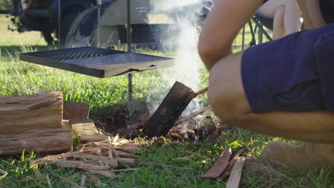Placing-wood-on-an-open-campfire-BBQ-at-a-camp-site-with-tent-and-vehicle-in-the-background