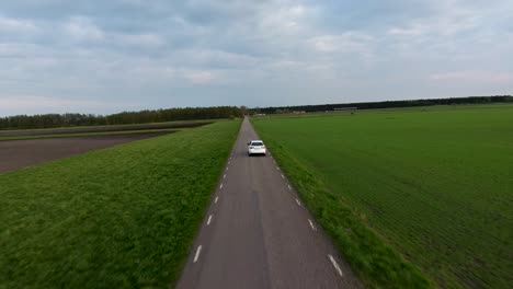 Amplia-Toma-Aérea-De-Un-Auto-Blanco-Conduciendo-Por-La-Estrecha-Carretera-Rural-En-El-Sur-De-Suecia-Skåne