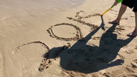 A-man-writing-"2018"-with-the-stick-end-of-the-shovel-on-a-sandy-beach-in-Oahu,-Hawaii