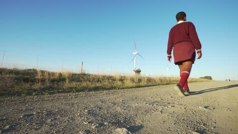 School-girl-walking-on-a-gravel-road-next-to-wind-turbine's,-low-angle
