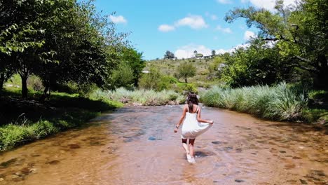 Slow-motion-flyover-drone-shot-of-a-barefooted-woman-in-a-white-dress-walking-happily-through-a-fresh-water-stream-in-the-middle-of-the-forest