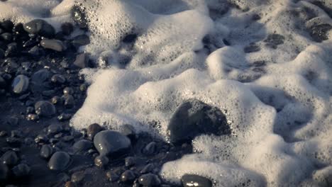Close-up,-slow-motion-view-of-waves-from-ocean-washing-over-wet-stones-on-beach-of-Tenerife-with-sunlight-shining-on-foaming-water