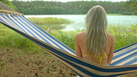 Young-beautiful-blonde-girl-sitting-and-swinging-on-blue-and-white-stripy-hammock-by-the-lake-looking-out-and-enjoying-the-view