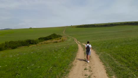 Aerial-follow-shot-of-male-walking-on-dirt-path-through-green-countryside-on-sunny-day