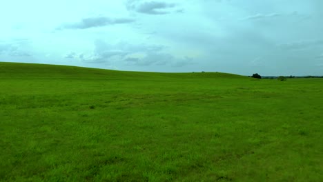 Vuelo-Lento-Sobre-El-Campo-De-Hierba-Verde,-Cielo-Azul-Y-Nubes-Grises,-Toma-Aérea,-Aspecto-Cinematográfico