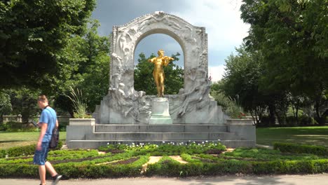 A-Man-Walking-In-Front-Of-The-Famous-Johann-Strauss-Monument-In-Stadtpark,-Vienna,-Austria-On-A-Sunny-Day---Medium-Shot
