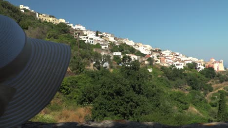 Right-pan-from-back-of-girl-with-summer-hat,-looking-out-on-Greek-town-on-hill-in-distance-SLOW-MOTIOn
