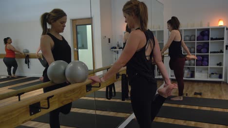 Damen-Trainieren-Vor-Dem-Spiegel-In-Einem-Yoga-Fitnessstudio,-Tragen-Trainingsanzüge,-An-Der-Ecke-Steht-Ein-Ständer-Mit-Trainingsbällen