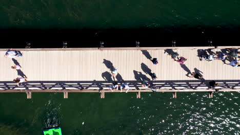 Luftaufnahme:-Ansicht-Von-Oben-Nach-Unten-Von-Menschen,-Die-Auf-Der-Holzbrücke-In-Trakai-Gehen,-Mit-Grün-Gefärbtem-Seewasser-Im-Hintergrund