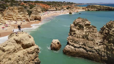 Fisherman-stands-atop-rocks-in-foreground,-Albufeira-Beach-in-background,-Portugal,-high-angle