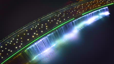 Aerial-left-to-right-top-down-tracking-view-of-Starlight-Bridge-or-Anh-Sao-Bridge-at-night,-a-pedestrian-bridge-with-colored-lights-and-waterfall-in-District-7-of-Ho-Chi-Minh-City-or-Saigon,-Vietnam