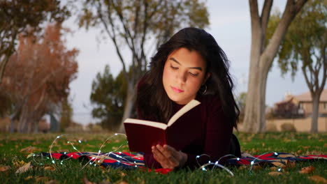A-cute-young-woman-reading-a-story-book-laying-in-the-park-at-twilight-in-autumn