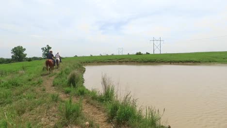 Two-cowboys-on-horseback-ride-away-from-a-waterhole,-Kansas