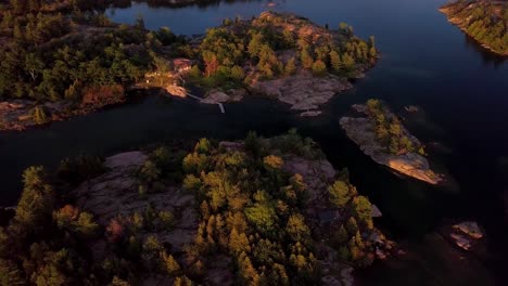 Rocky-Islands-with-Abandoned-Cabin-and-Pine-Trees-in-Blue-Lake-at-Sunset,-Drone-Aerial-Wide-Pedestal-Up-Tilt-Down-Overhead