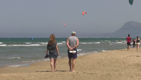 Middle-aged-couple-walking-on-sandy-beach-with-kitesurfers-in-background,-wide-shot