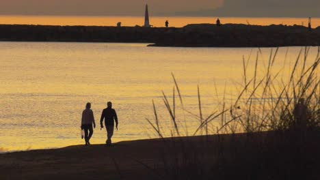 Older-couple-walking-on-beach-together-at-dawn,-silhouette-against-calm-sea,-slow-motion