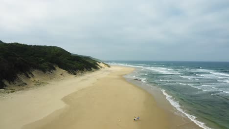 Aerial,-family-walks-on-huge-sandy-beach-with-ocean-waves,-cloudy-sky