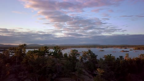 Fly-over-a-Rocky-Pine-Tree-Hill-to-Reveal-Small-Granite-Islands,-Blue-Sky-and-Clouds-at-Sunset,-Drone-Aerial-Wide-Dolly-In