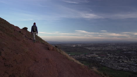 Dolly-tracking-shot-of-girl-walking-up-the-Arthurs-seat-mountain-on-the-hiking-trail-in-evening,-dusk-with-city-of-Edinburgh-in-the-background
