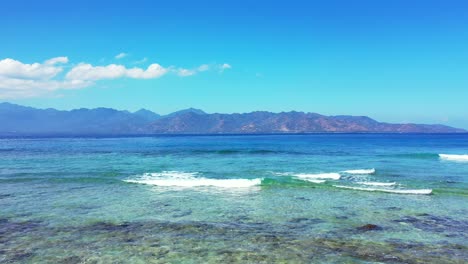 Beautiful-seashore-with-shallow-calm-water-over-coral-reefs-and-rocky-seabed-where-white-waves-splashing-and-foaming-with-blue-sea-and-mountains-background