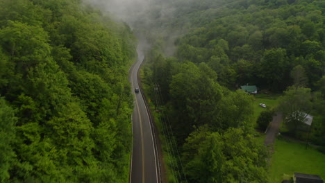 Aerial-drone-overhead-of-single-car-driving-down-curvy-mountain-road-through-green-forest-valley-with-trees-and-rainy-fog
