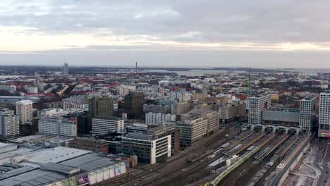 Aerial,-pan,-drone-shot-over-looking-the-cityscape,-trains-and-traffic,-revealing-the-Mall-of-Tripla-shopping-center,-and-the-Pasila-railway-station,-on-a-cloudy-evening,-in-Helsinki,-Finland