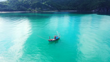 Fishing-boat-floating-on-calm-turquoise-lagoon-near-coastline-of-tropical-island-with-rocks-and-beaches-of-green-hills