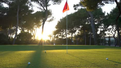 Ein-Golfball-Fliegt-Schnell-An-Der-Flagge-Vorbei,-Die-Rot-Ist-Und-Bei-Sonnenuntergang-Im-Wind-Flattert.-Eine-Wunderschöne-Sonne-Beleuchtet-Den-Hintergrund