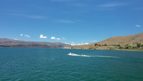 Eine-Drohnenaufnahme-Eines-Mannes-Und-Eines-Jungen,-Die-Auf-Einem-Aufblasbaren-Schlauch-Hinter-Einem-Schnellboot-Im-Zickzack-Auf-Dem-Wunderschönen-Blauen-Lake-Aviemore-In-Neuseeland-Geschleppt-Werden