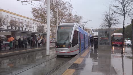 Light-rail-tram-leaving-train-stop-on-rainy-day-in-Seville,-Spain