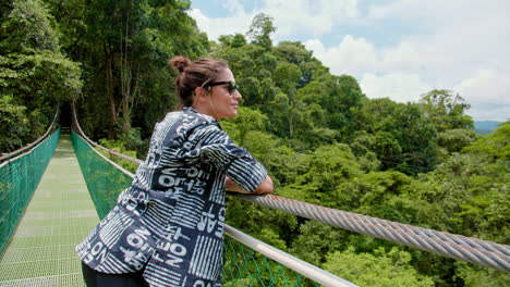 Moving-In-Shot-of-Beautiful-Brunette-Girl-with-Sunglasses-Enjoying-the-Panorama-on-Hanging-Bridge-in-the-Middle-of-the-Forest-in-Arenal,-Costa-Rica