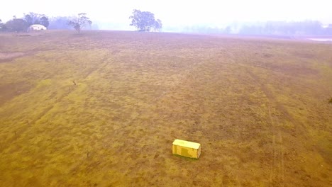 Abandoned-trailer-box-near-bush-fire-with-heavy-smoke-in-the-distance,-drone-tilt-up-advance-view