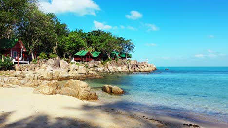 Calm-exotic-beach-with-sand-and-cliffs-on-shore-washed-by-calm-turquoise-sea,-bungalows-under-trees-with-ocean-view-in-Thailand