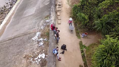 Vietnamese-woman-with-face-covered-moving-a-wheelbarrow-with-gravel-passing-some-male-tourists,-Top-view-pan-down-shot
