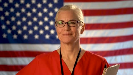 Mittelscharfes-Porträt-Einer-Krankenschwester-Mit-Brille,-Die-Glücklich-Auf-Die-Kamera-Zugeht,-Vor-Einem-Unscharfen-Hintergrund-Mit-Amerikanischer-Flagge